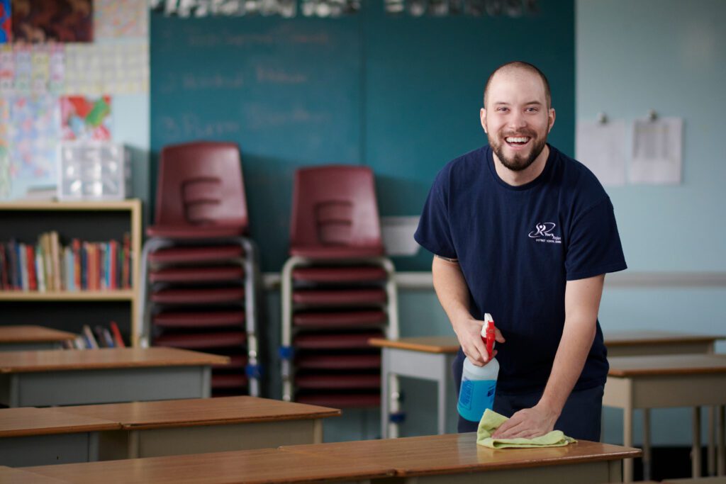 Highlights - Tara Mansfield Portfolio. Image of a custodian smiling at the camera holding a cloth and cleaning spray.