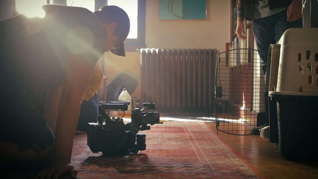 Image of videographer kneeling on floor with DJI Ronin 4D camera pointed towards large dog crate where a collie's nose pokes out of.