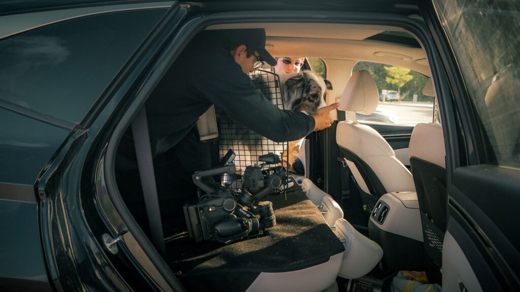 Videographer in black sweater with DJI Ronin 4D camera inside back of car giving treat to white, brown and grey collie dog